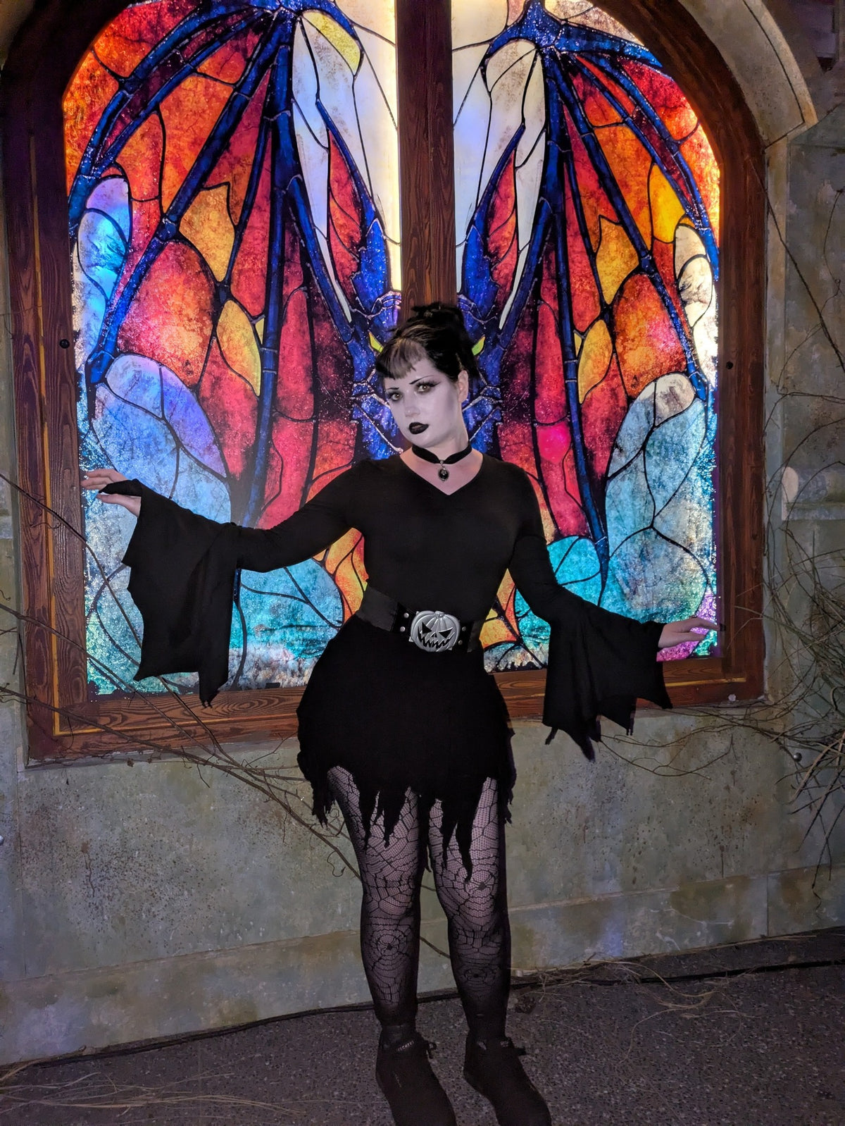 Ren is wearing the black pumpkin waist belt with a bat-sleeve dress and spiderweb tights in front of colorful stained glass.