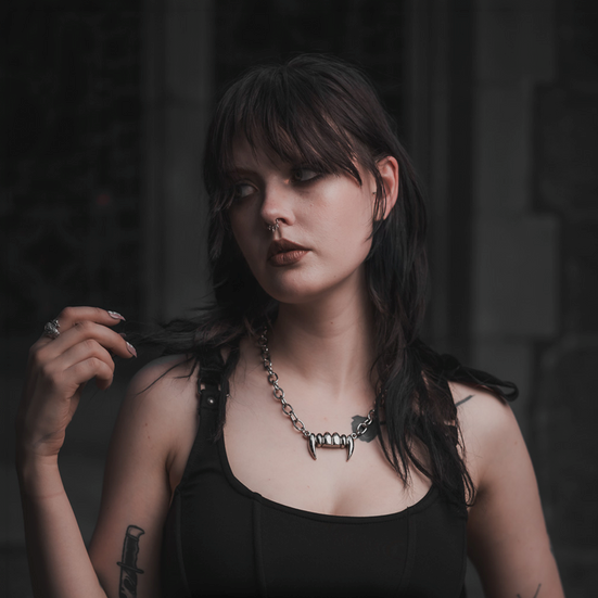 Goth woman with dark hair and piercings wearing a black tank top and our vampire fang chain necklace in a dimly lit setting.