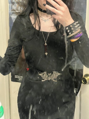 A goth woman taking a mirror selfie wearing a black long-sleeve dress with a square neckline, a silver bat-belt, and layered necklaces, standing in front of a door.  Snag the silver bat belt in our store.