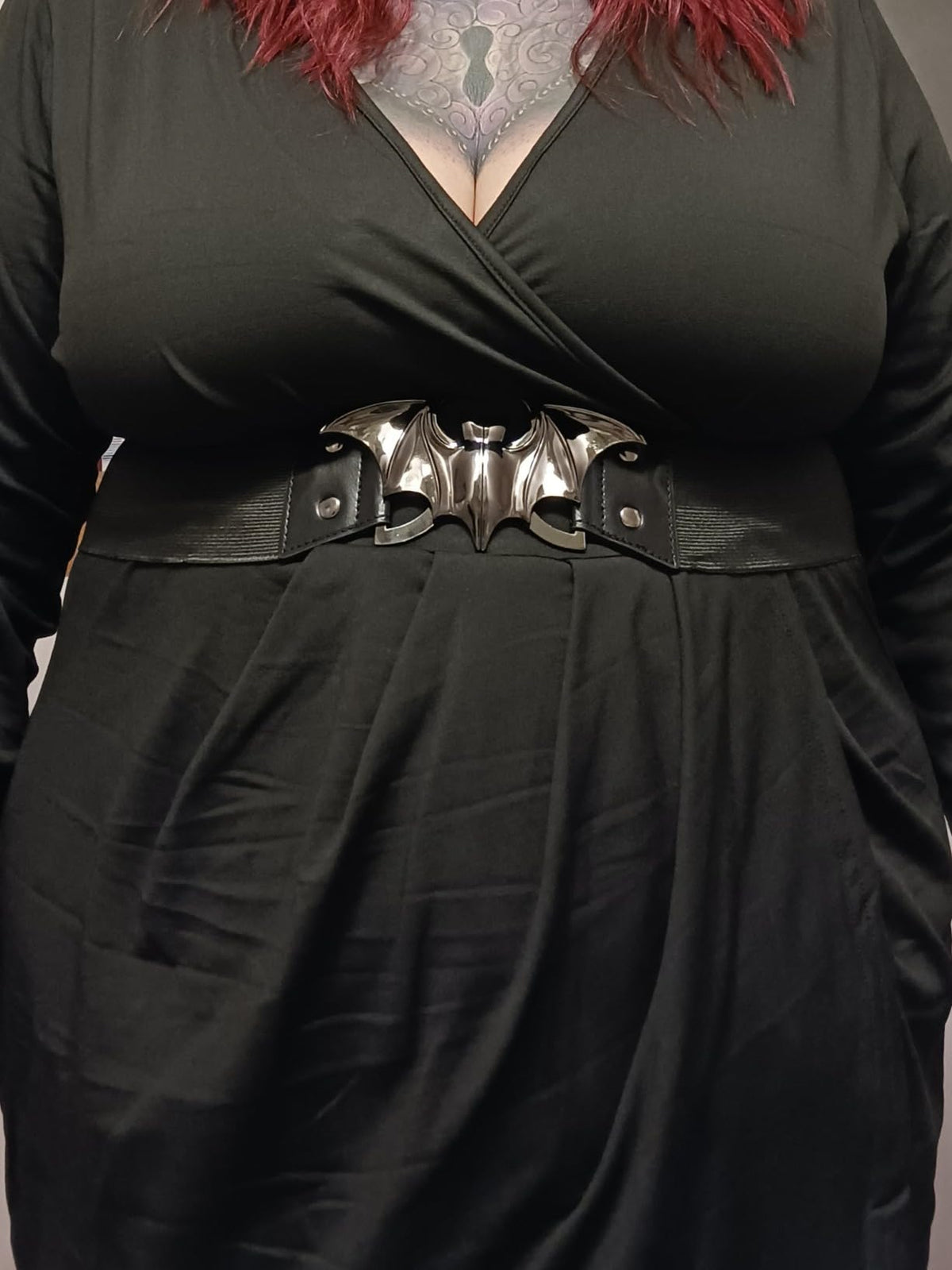 A close-up of a goth woman wearing our black wrap-style dress with a wide black belt featuring a metallic silver bat-shaped centerpiece.