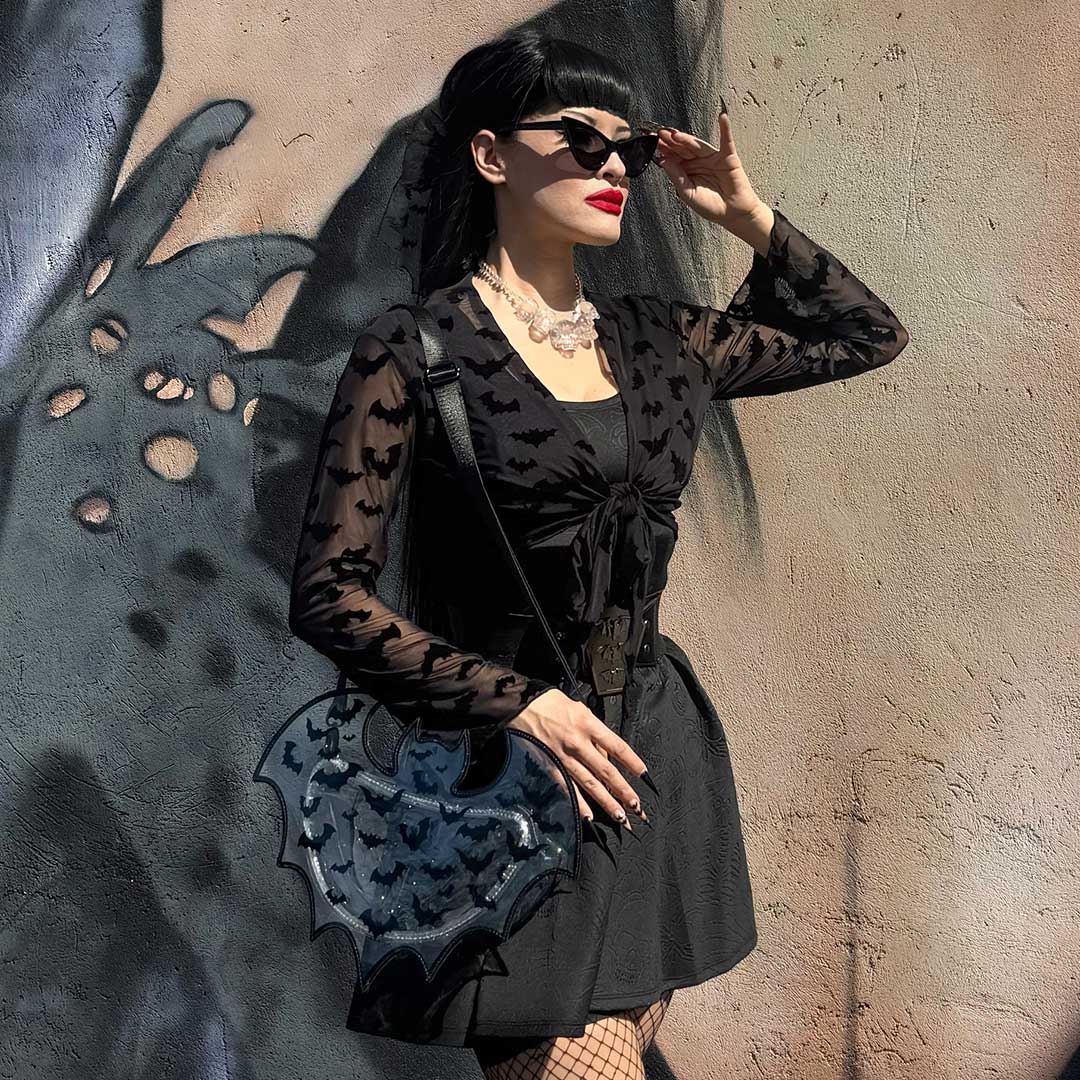 Woman in a black dress with bat patterns holding a matching bag against a textured wall.