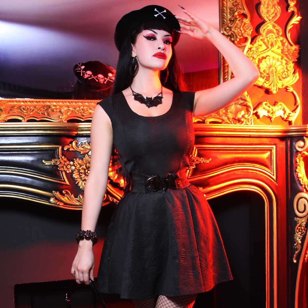 A woman posing in a black gothic-style dress with a flared skirt, matching black hat with a cross, and accessories, set against an ornate red and gold fireplace.